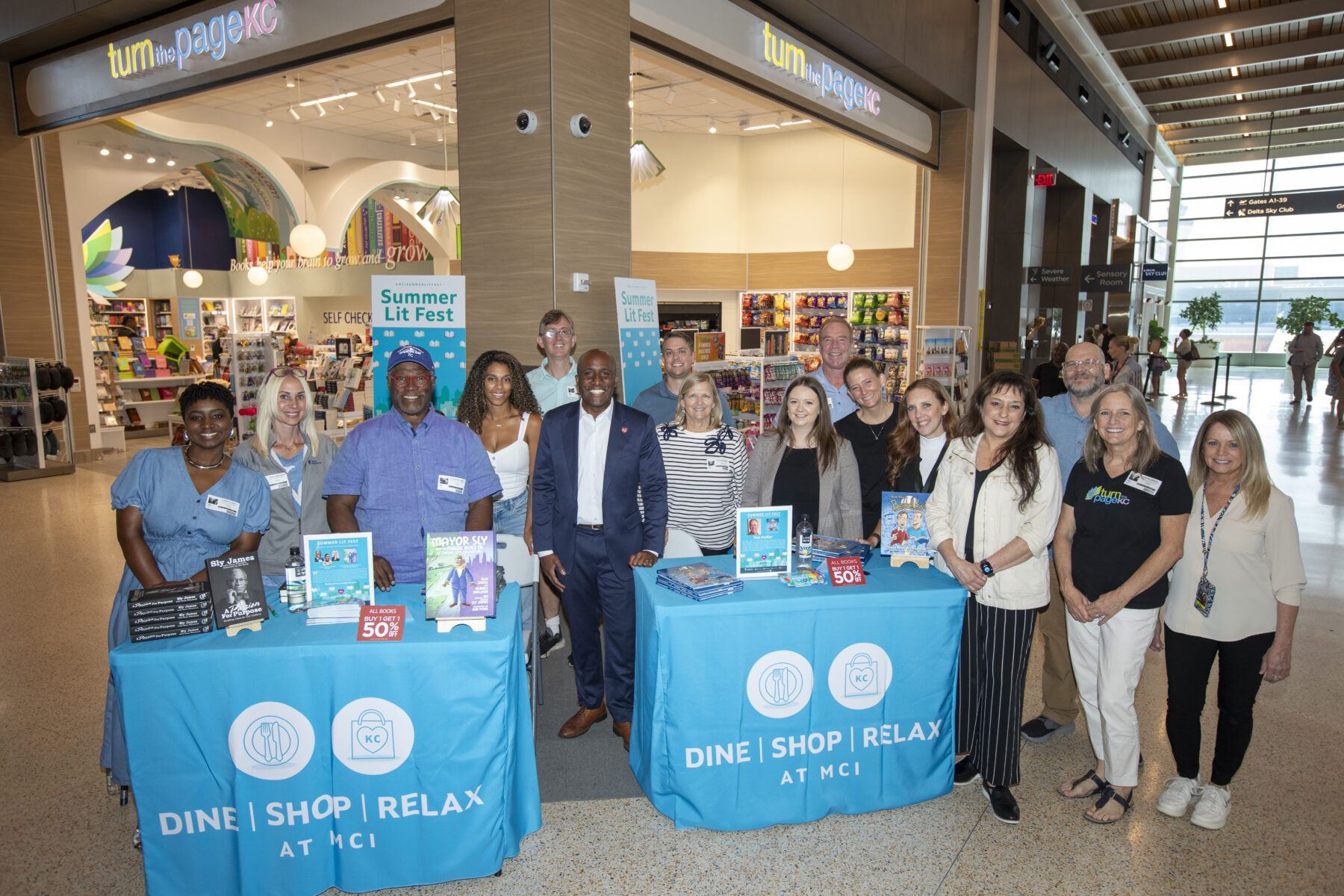 A group of community members and event participants gathered inside an airport terminal in front of the Turn the Page KC bookstore during a Summer Lit Fest event. Two promotional tables with books, event materials, and “Dine | Shop | Relax at MCI” branding are displayed in the foreground. Store signage, bookshelves, and airport concourse wayfinding signs are visible in the background.