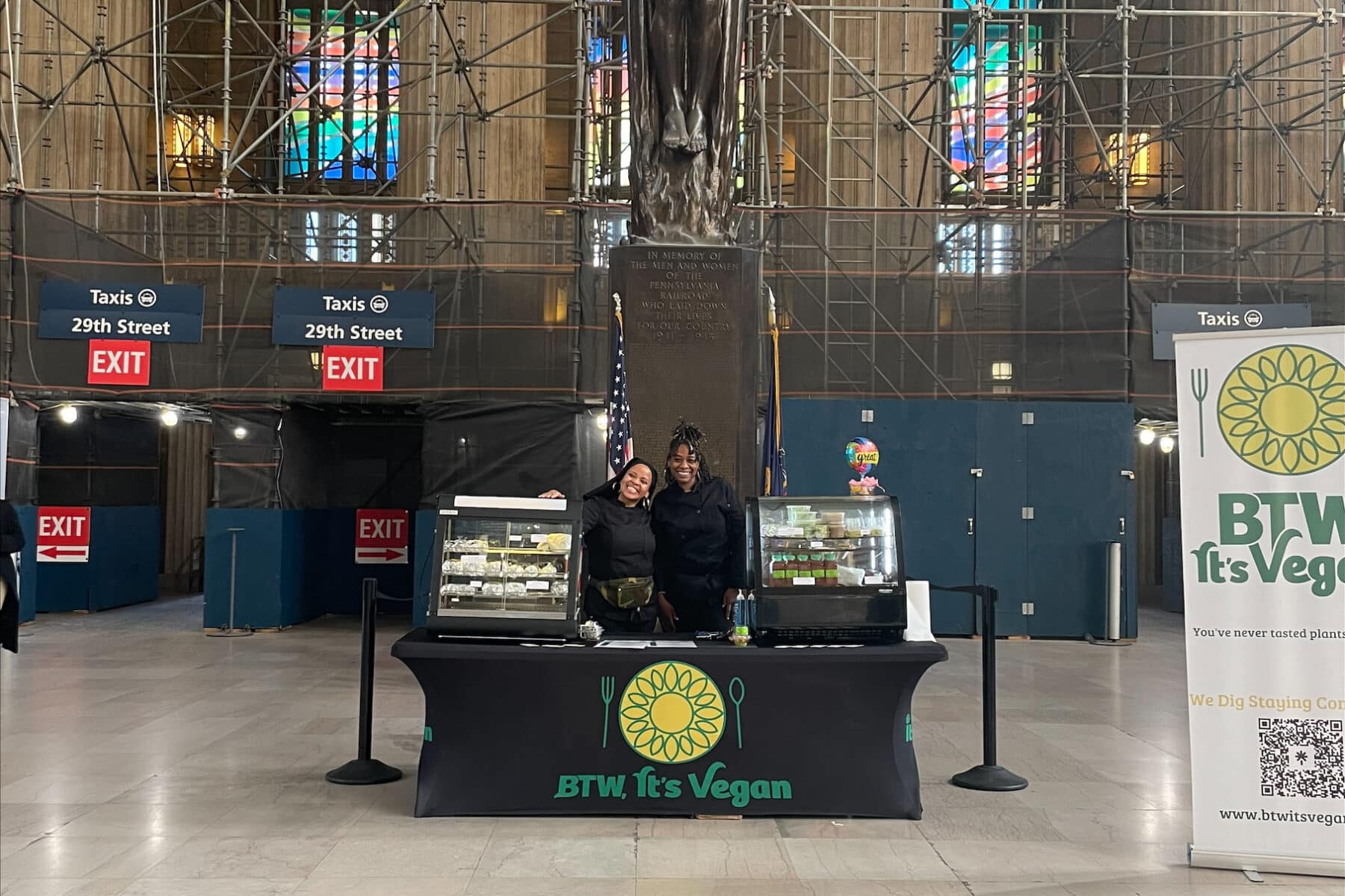 A vendor display for BTW, It’s Vegan is set up inside a large transportation terminal. Two staff members stand behind a branded table featuring packaged vegan food items in refrigerated display cases. The space includes high ceilings, stained-glass windows, directional signs for taxis and exits, and scaffolding surrounding a central monument in the background. A promotional banner for the vegan food business is displayed beside the booth.