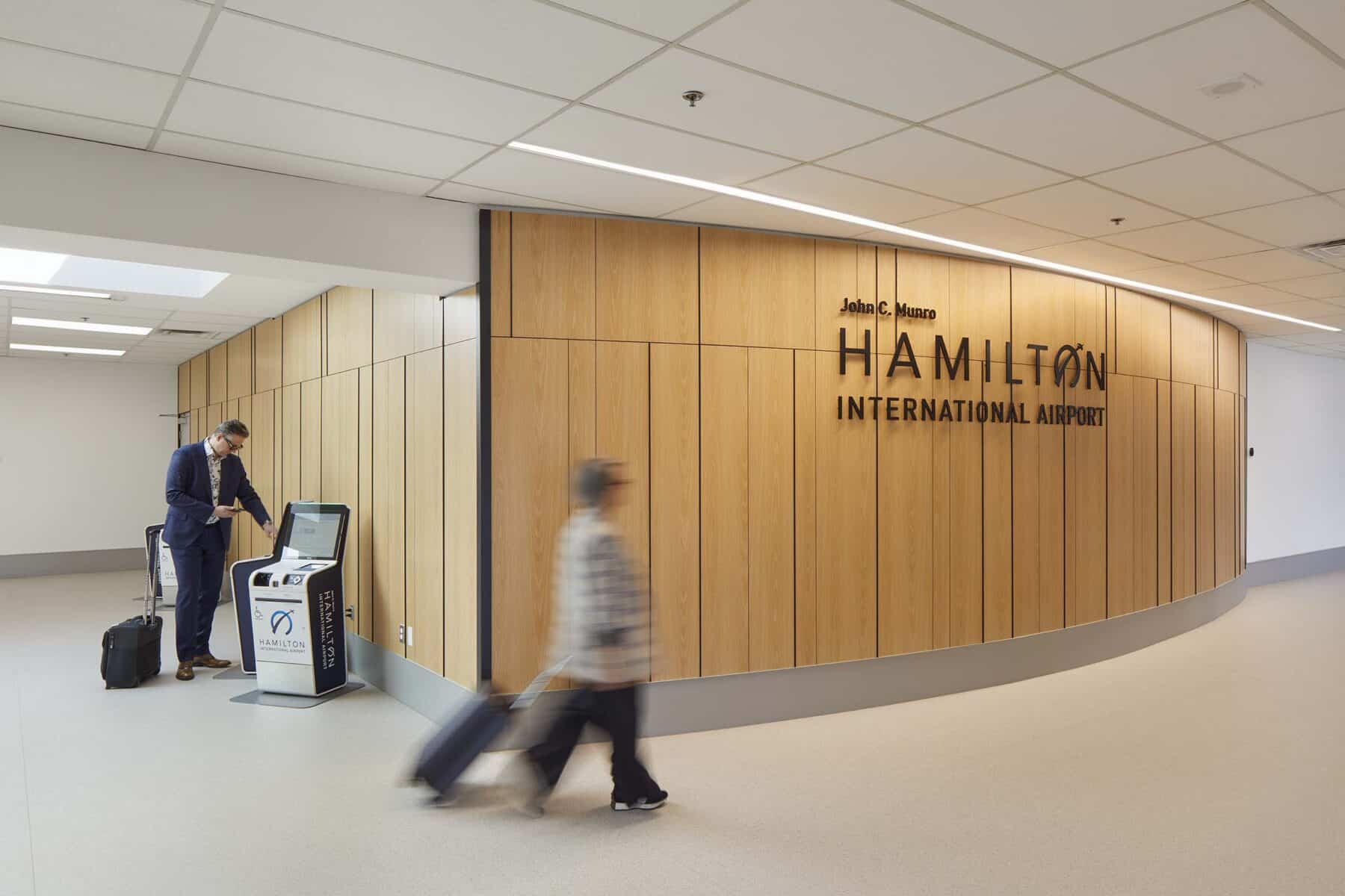 A traveler walks through a corridor at John C. Munro Hamilton International Airport, passing a wood-paneled wall with the airport’s name displayed in large lettering, while another traveler uses a self‑service kiosk nearby.