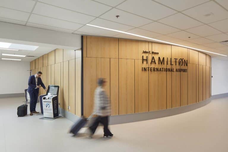 A traveler walks through a corridor at John C. Munro Hamilton International Airport, passing a wood-paneled wall with the airport’s name displayed in large lettering, while another traveler uses a self‑service kiosk nearby.