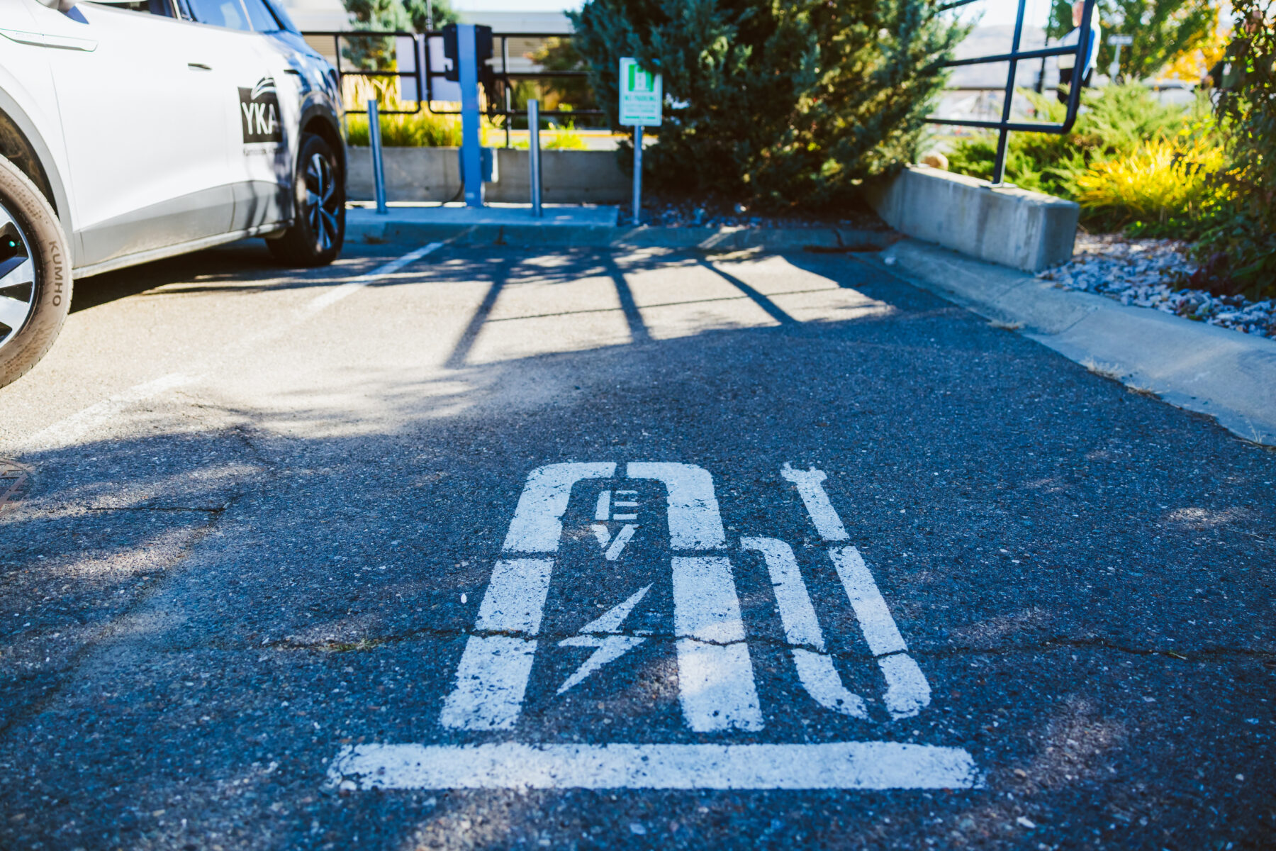An electric vehicle charging station in an airport parking area, with a white airport-branded vehicle parked beside it. A painted EV charging symbol is visible on the asphalt in the foreground, while the charging equipment, landscaping, and railings are seen in the background under bright daylight.