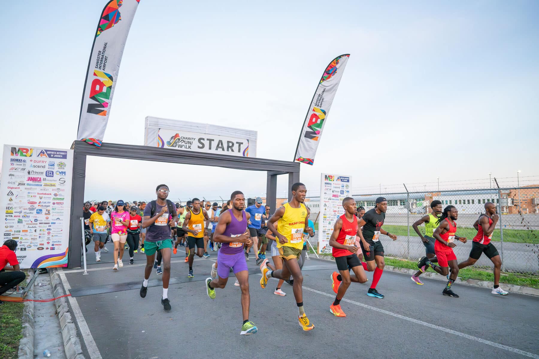 Participants begin the MBJ Airports Charity Run/Walk, crossing the start line at Sangster International Airport in Jamaica. Runners in colorful athletic gear move down the roadway beneath a large “START” banner, with event flags, sponsor signage, airport facilities, and perimeter fencing visible in the background.