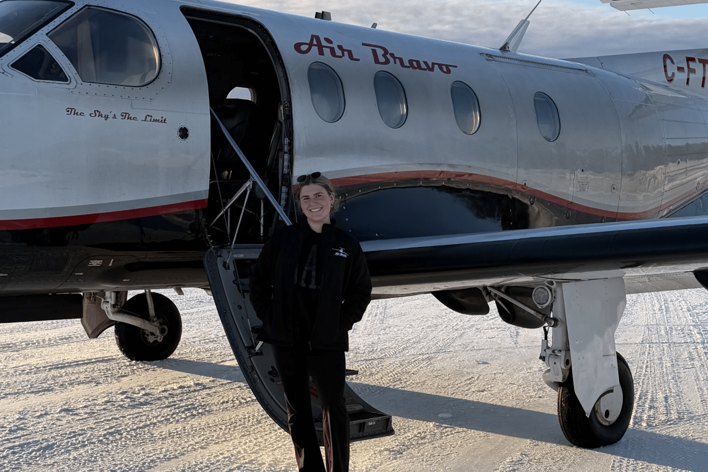 A person stands beside the open door of a small Air Bravo aircraft parked on a snowy airport apron. The white aircraft with red and black trim features the words “Air Bravo” on the fuselage and “The Sky’s The Limit” near the cockpit. The person is positioned beneath the aircraft wing, with the open entry stairs visible and winter sunlight reflecting off the snow-covered ground.