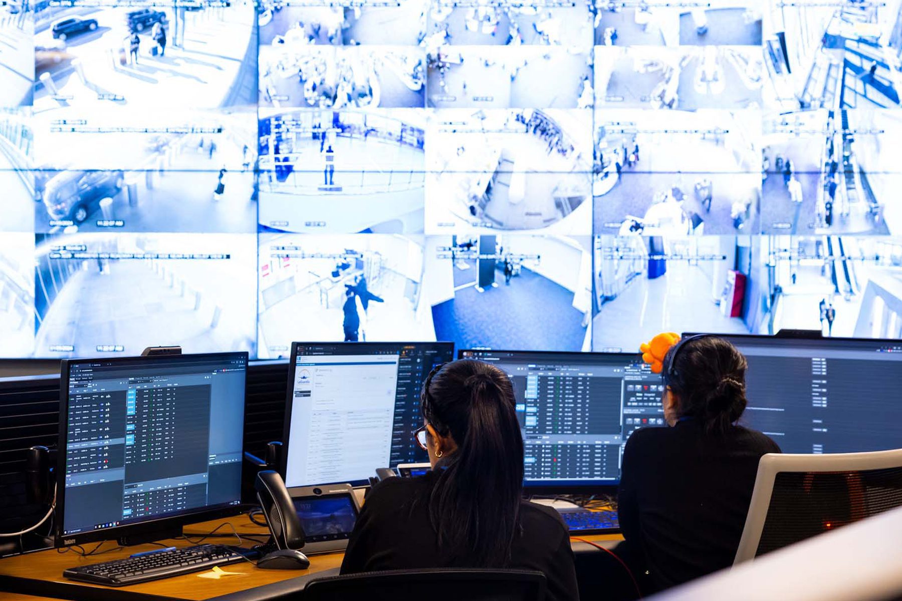Two people sit in a room with a large screen showing various security camera footage at an airport terminal.