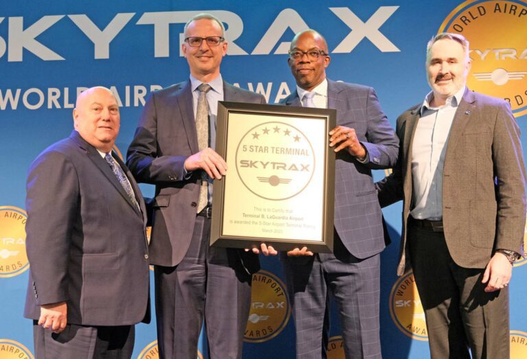Four men in suits pose with a large award that reads "5 star terminal, SkyTrax" awarded to the Terminal B of LaGuardia Airport.
