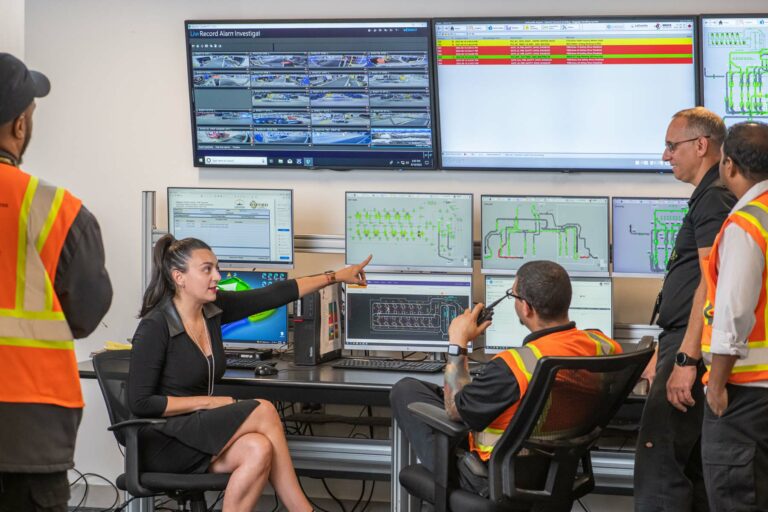 A group of airport employees point and discuss the layout of a terminal on computer screens at air traffic control.