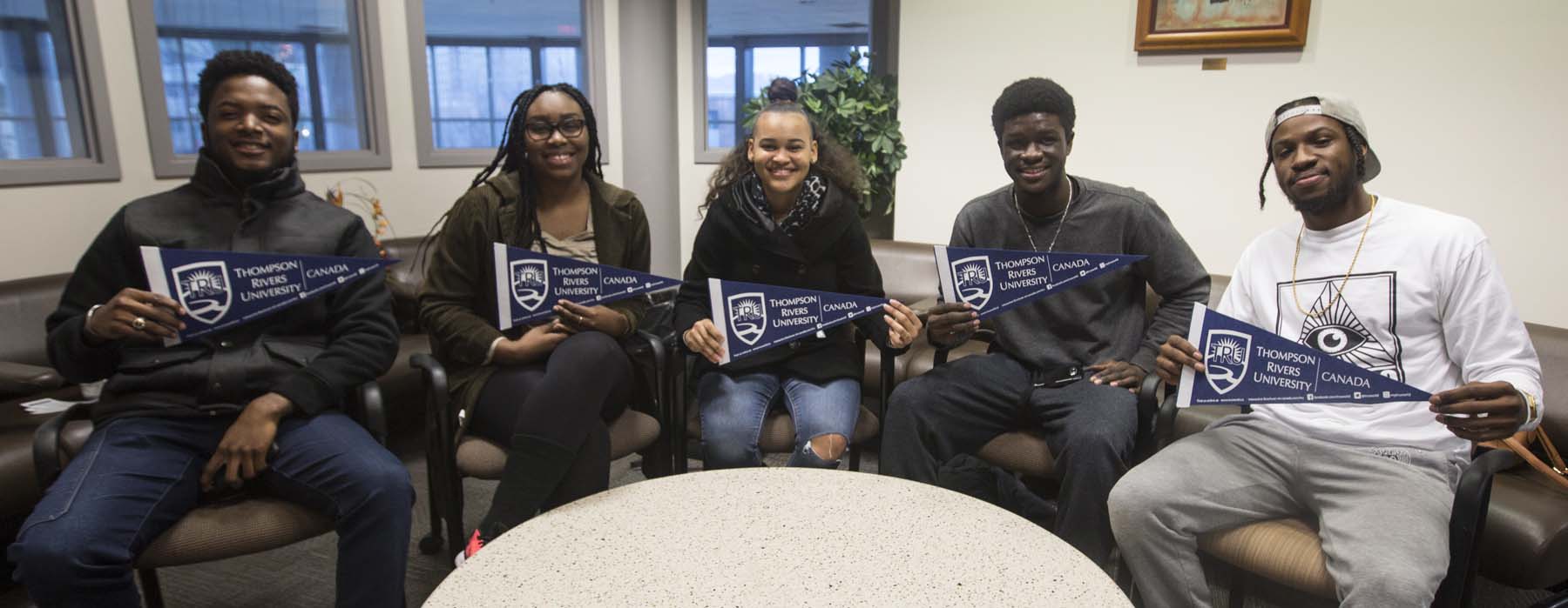 Five people hold up navy blue Thompson Rivers University banners while sitting and smiling for the camera.