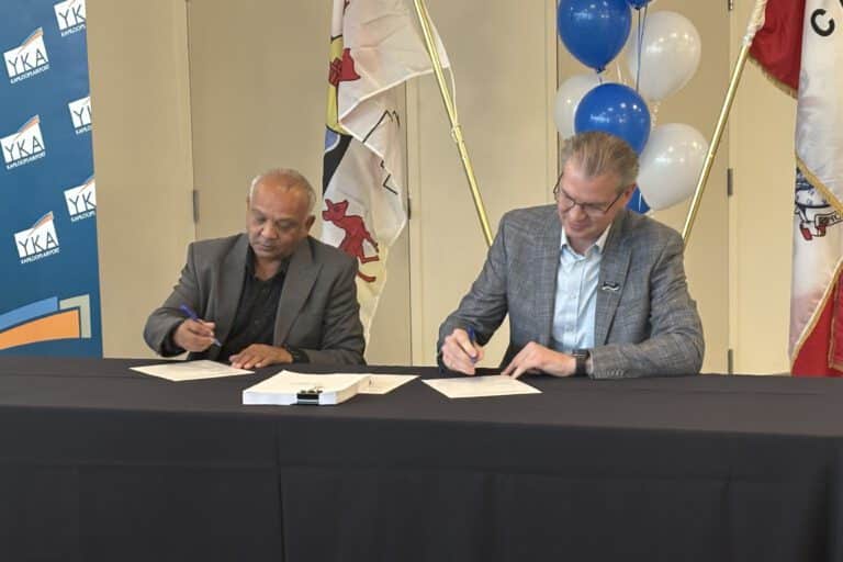 Two men sit at a table signing documents. Behind them are banners, balloons, and flags.