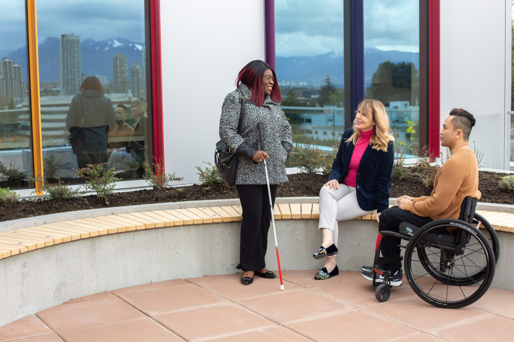 Three people, including one with a cane and another in a wheelchair, are talking on a circular bench outdoors with city buildings and mountains in the background.