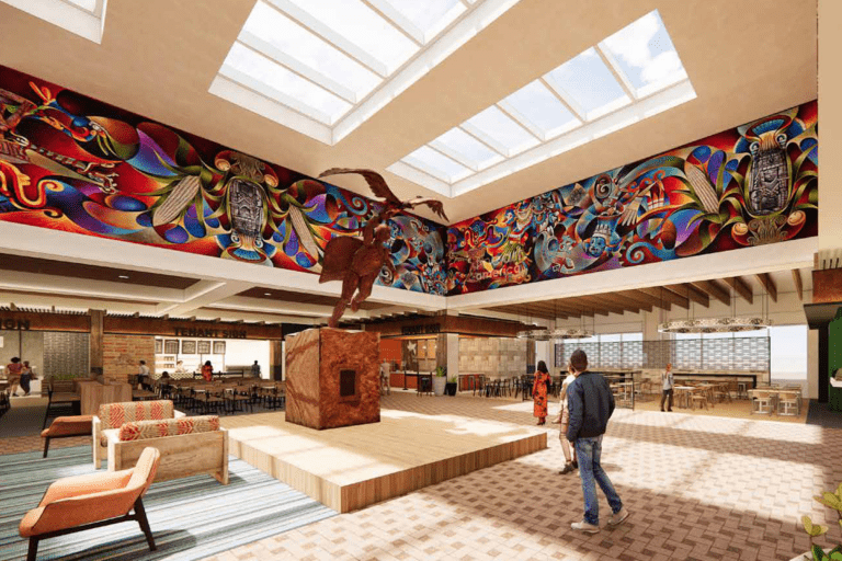 Spacious room with colorful mural and skylight, featuring a central statue on a wooden platform. People walk around, and there are seating areas and tables in the background.