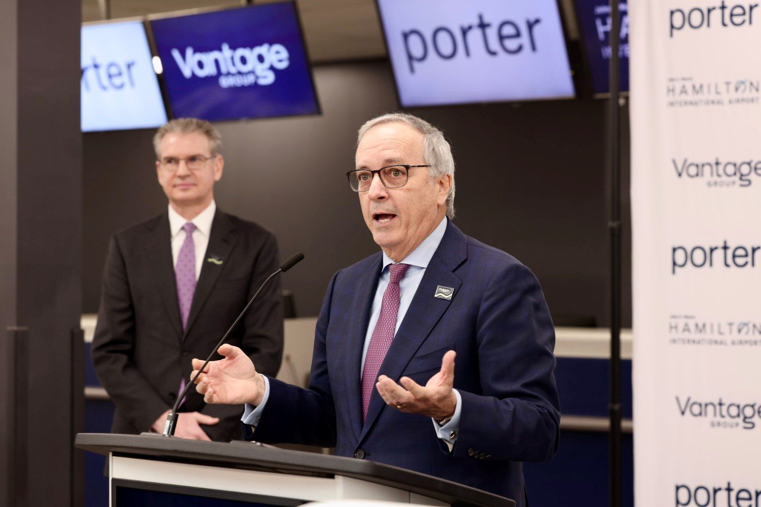 A man in a suit speaks at a podium with "porter" and "Vantage" signs in the background. Another man stands behind him.