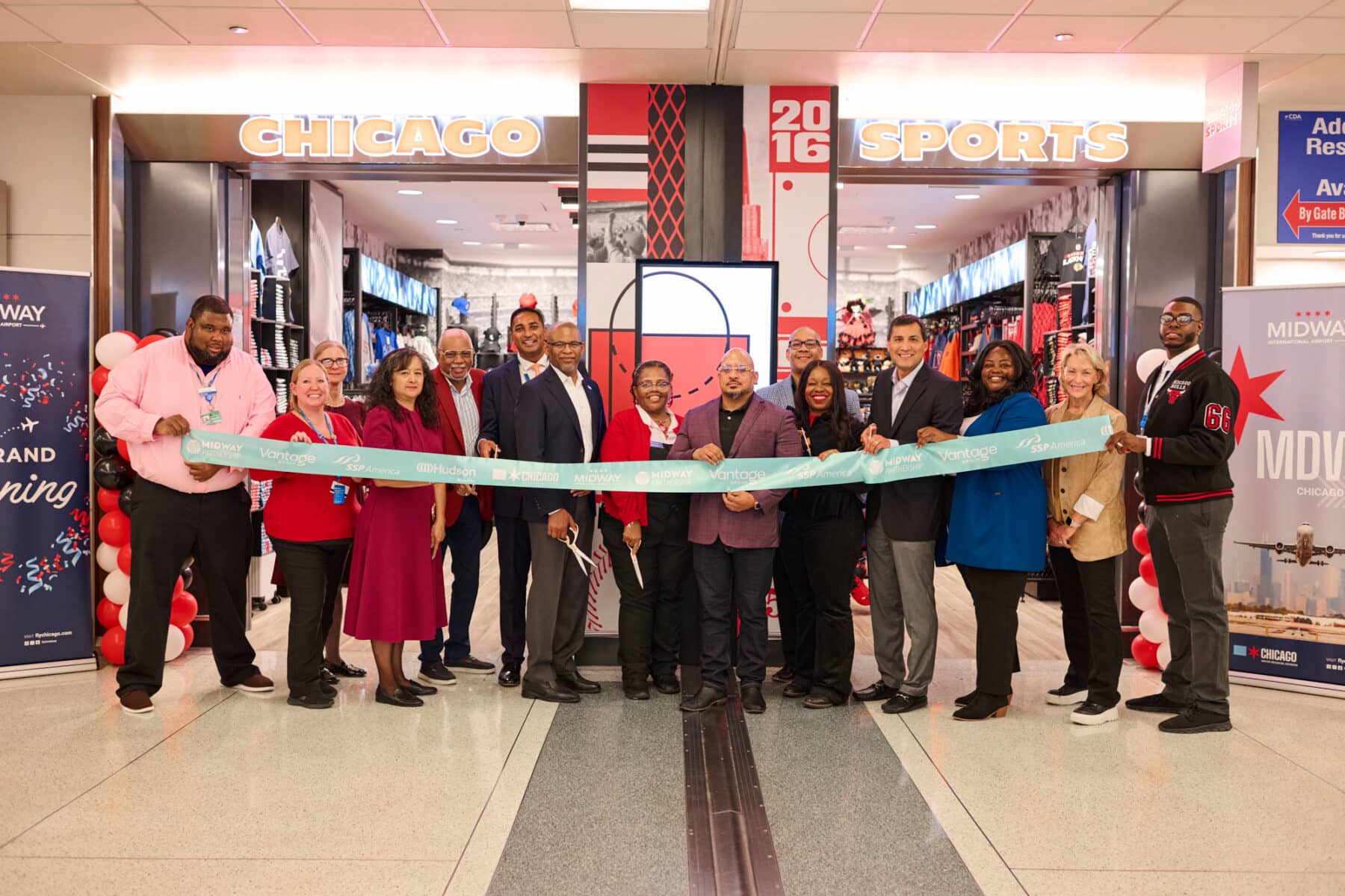 Ribbon-cutting ceremony at the new Chicago Sports store in Midway International Airport, with attendees holding a blue ribbon in front of the entrance decorated with red and white balloons and visible sports merchandise inside.