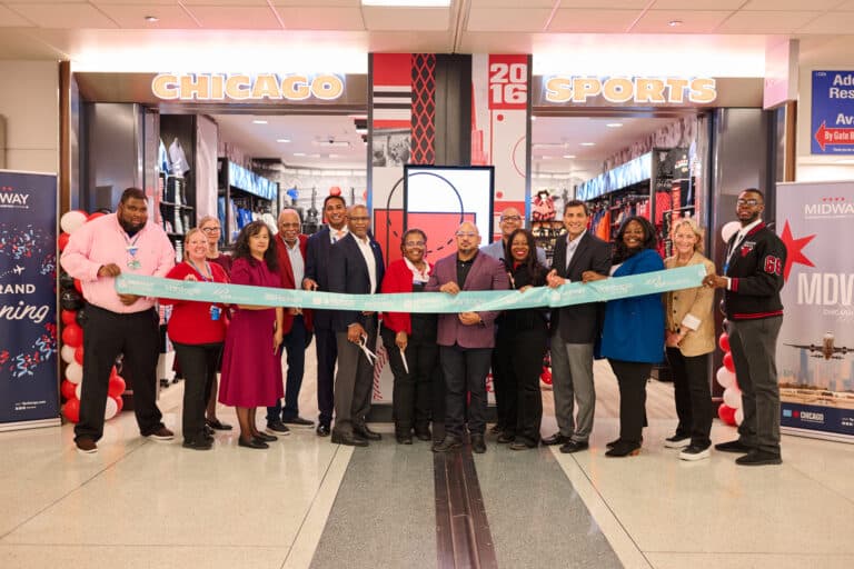 Ribbon-cutting ceremony at the new Chicago Sports store in Midway International Airport, with attendees holding a blue ribbon in front of the entrance decorated with red and white balloons and visible sports merchandise inside.
