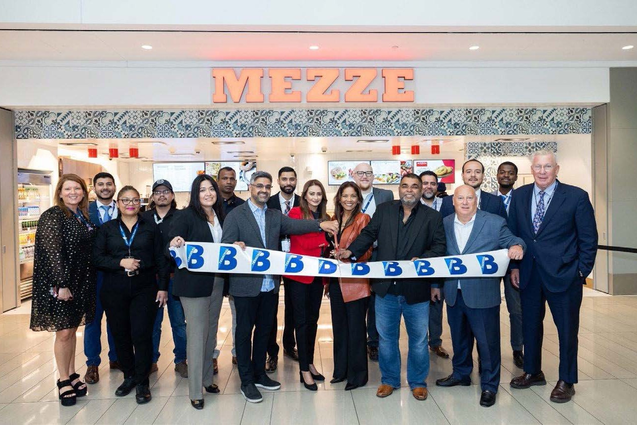 A group of people stand in front of a restaurant named 'MEZZE' during a ribbon-cutting ceremony at LaGuardia Terminal B. They hold a ribbon with the letter 'B' printed repeatedly. The restaurant features a modern design with patterned tiles above the entrance and visible food displays inside.