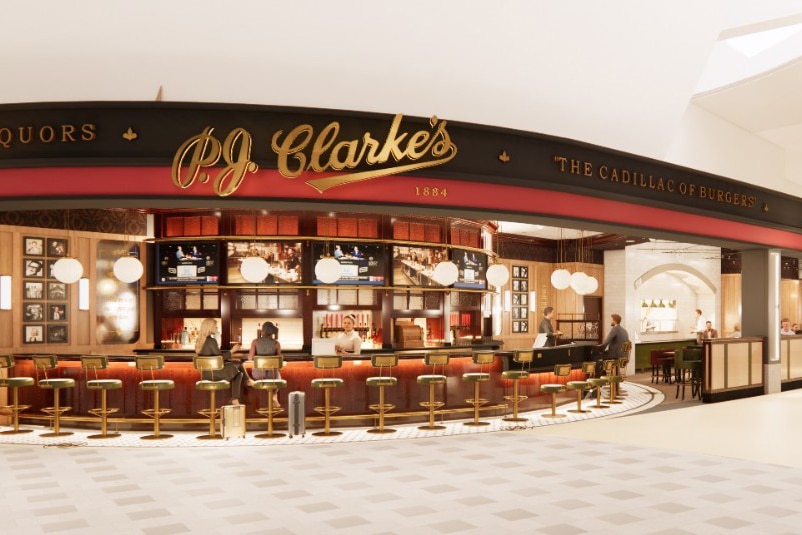 Front view of P.J. Clarke’s restaurant at JFK Terminal 6, featuring a classic bar with gold stools, warm lighting, and patrons seated at the counter under round ceiling lights.
