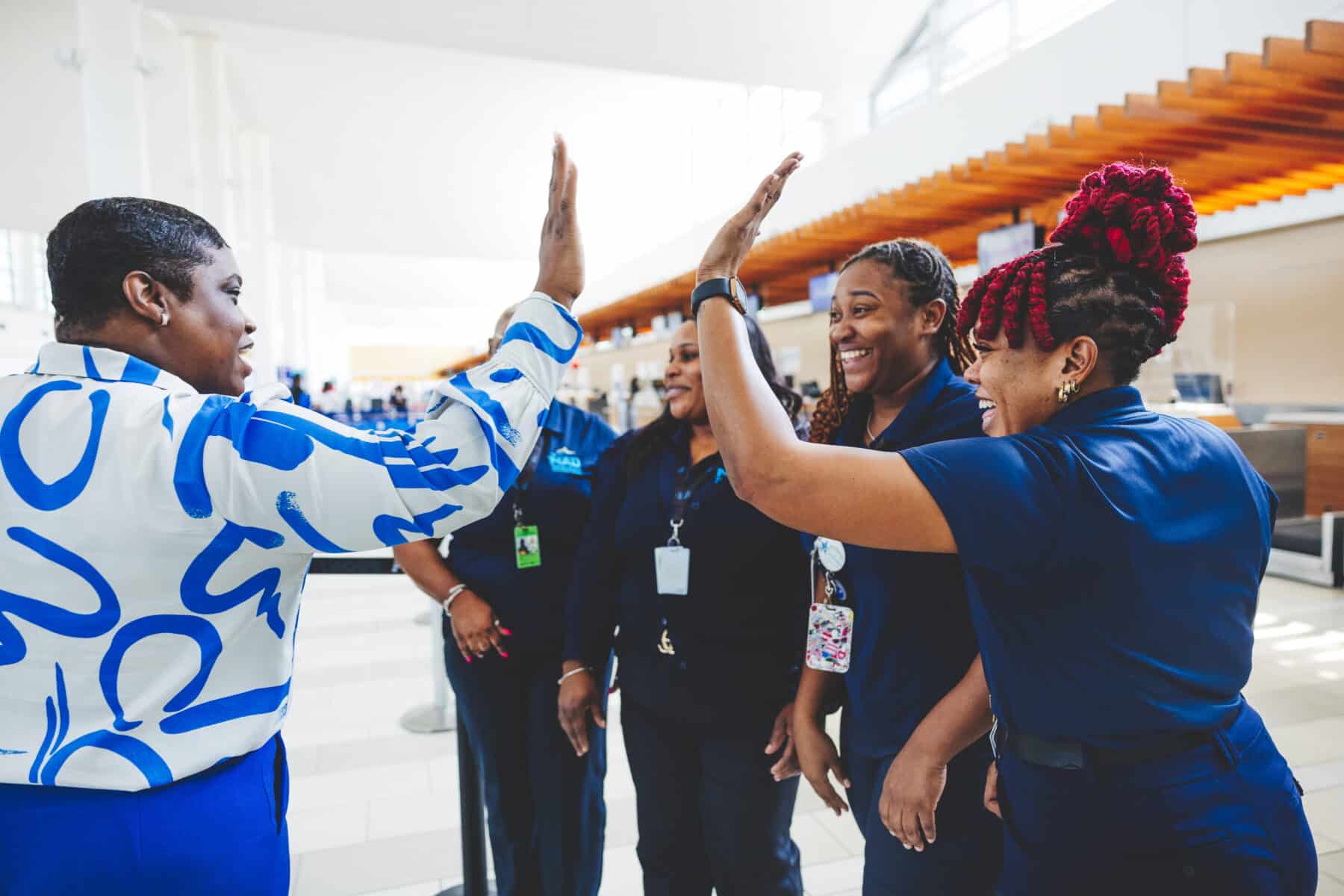 Group of uniformed employees standing together inside a modern, bright building with high ceilings and large windows. Two individuals in the foreground are giving each other a high-five, while others stand nearby wearing lanyards and ID badges.