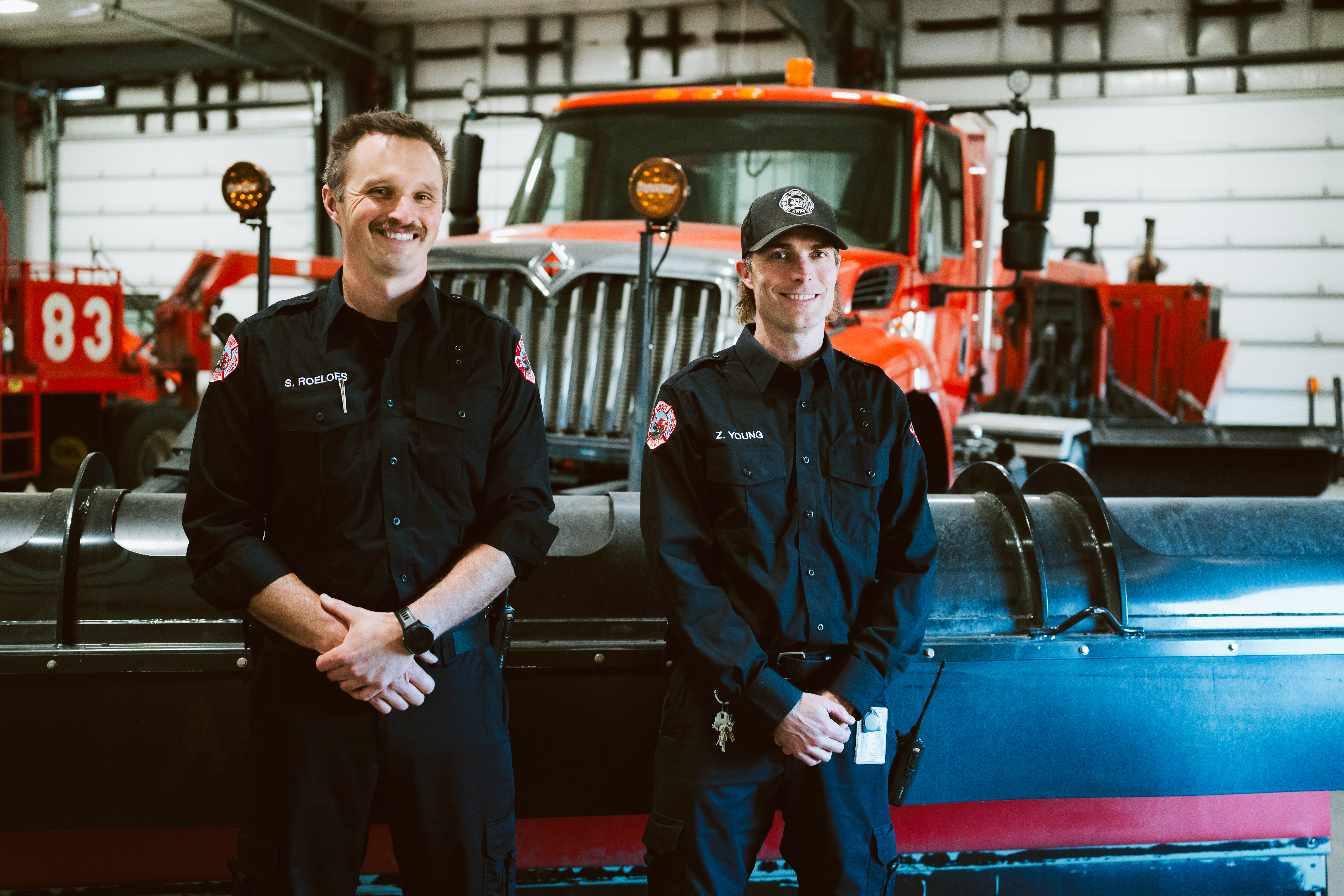wo uniformed firefighters standing indoors in front of a large red fire truck equipped with a snowplow attachment. The setting appears to be a fire station with garage doors and additional emergency vehicles visible in the background.