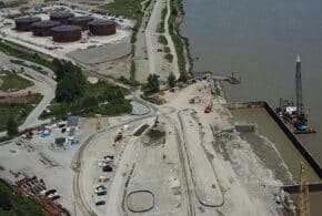 Aerial view of an industrial construction site near a river, featuring large cylindrical storage tanks, heavy machinery, and access roads.