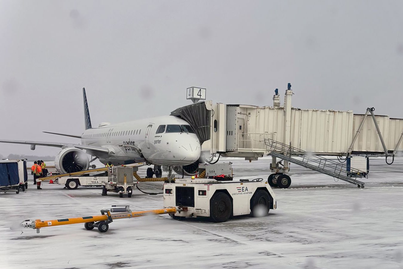 A passenger aircraft parked on a snowy tarmac is connected to a jet bridge, with ground crew and equipment working around the plane during winter weather.
