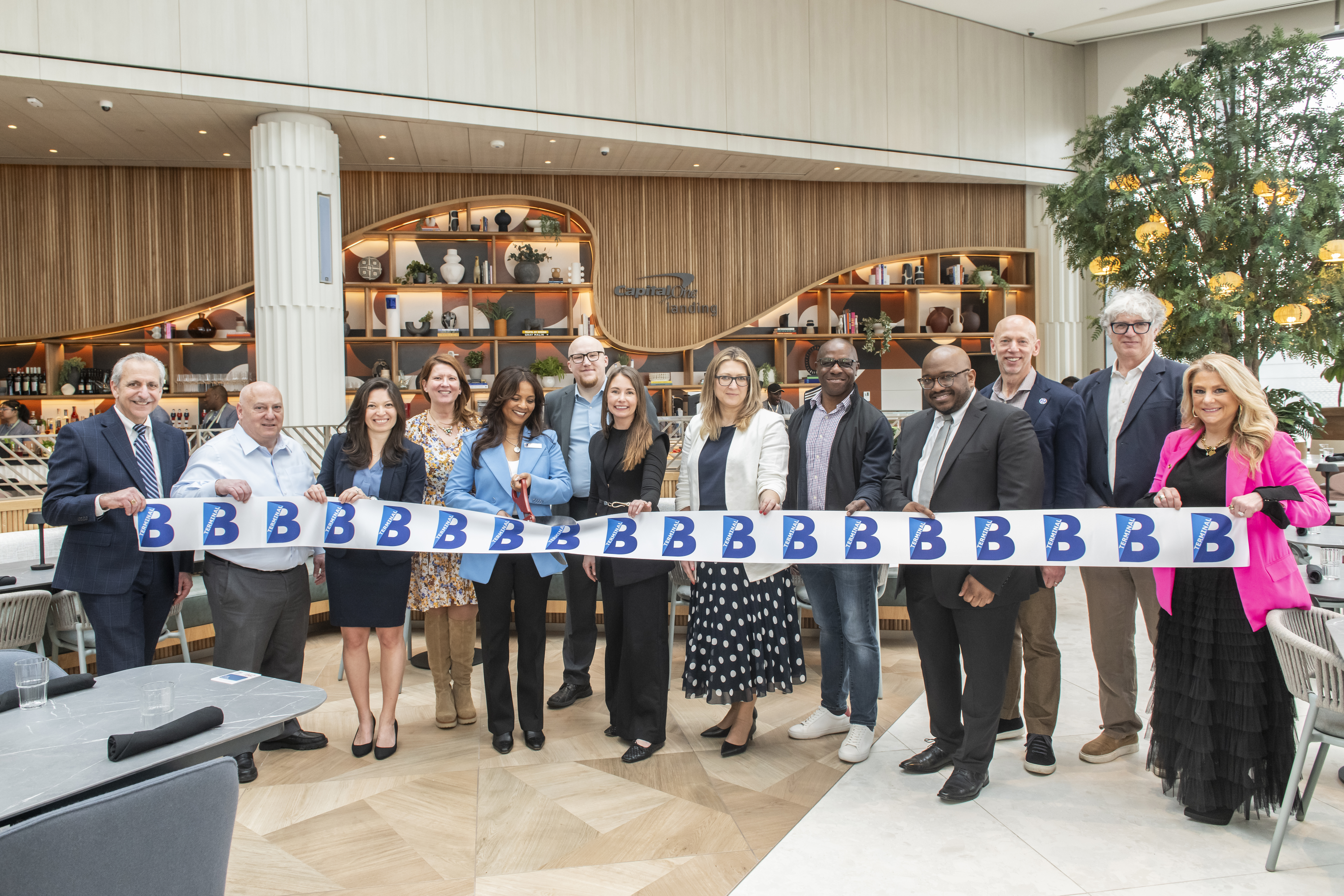 A group of people stand together in a bright, modern dining space, holding a ribbon decorated with a series of “B” logos during a ribbon‑cutting ceremony. Shelving with bottles and décor elements lines the wall behind them, and a large indoor tree is visible on the right side of the image.