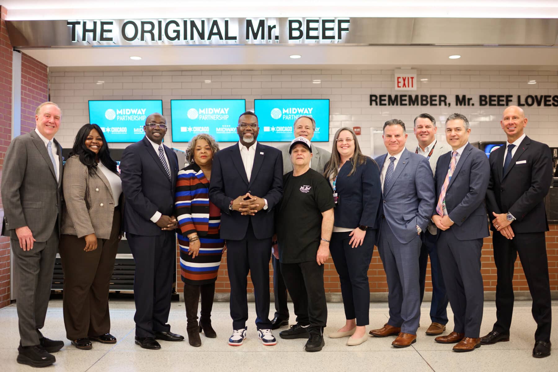 A group of people standing together in front of “The Original Mr. Beef” storefront at Chicago Midway International Airport, posing beneath the bright sign with digital Midway Partnership screens in the background. They are dressed in business and professional attire, marking the opening celebration of the new location.