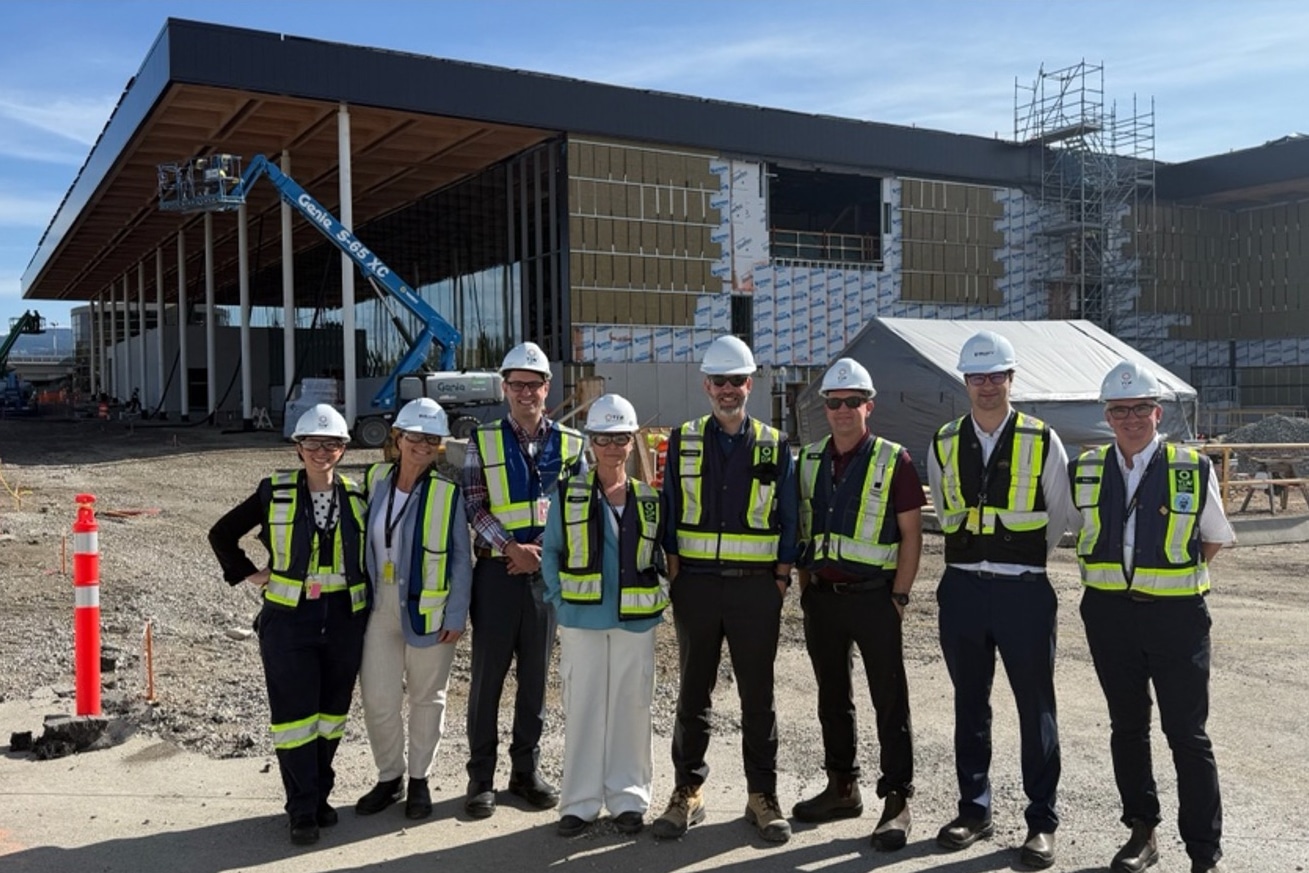 Project team wearing safety gear stands on site in front of a modern airport facility under construction, highlighting progress, collaboration, and infrastructure development.
