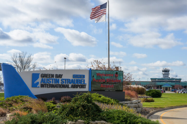 Entrance sign for Green Bay Austin Straubel International Airport (GRB) surrounded by landscaped greenery, with a digital welcome message, the U.S. flag flying overhead, and the airport terminal and control tower visible in the background under a partly cloudy sky.