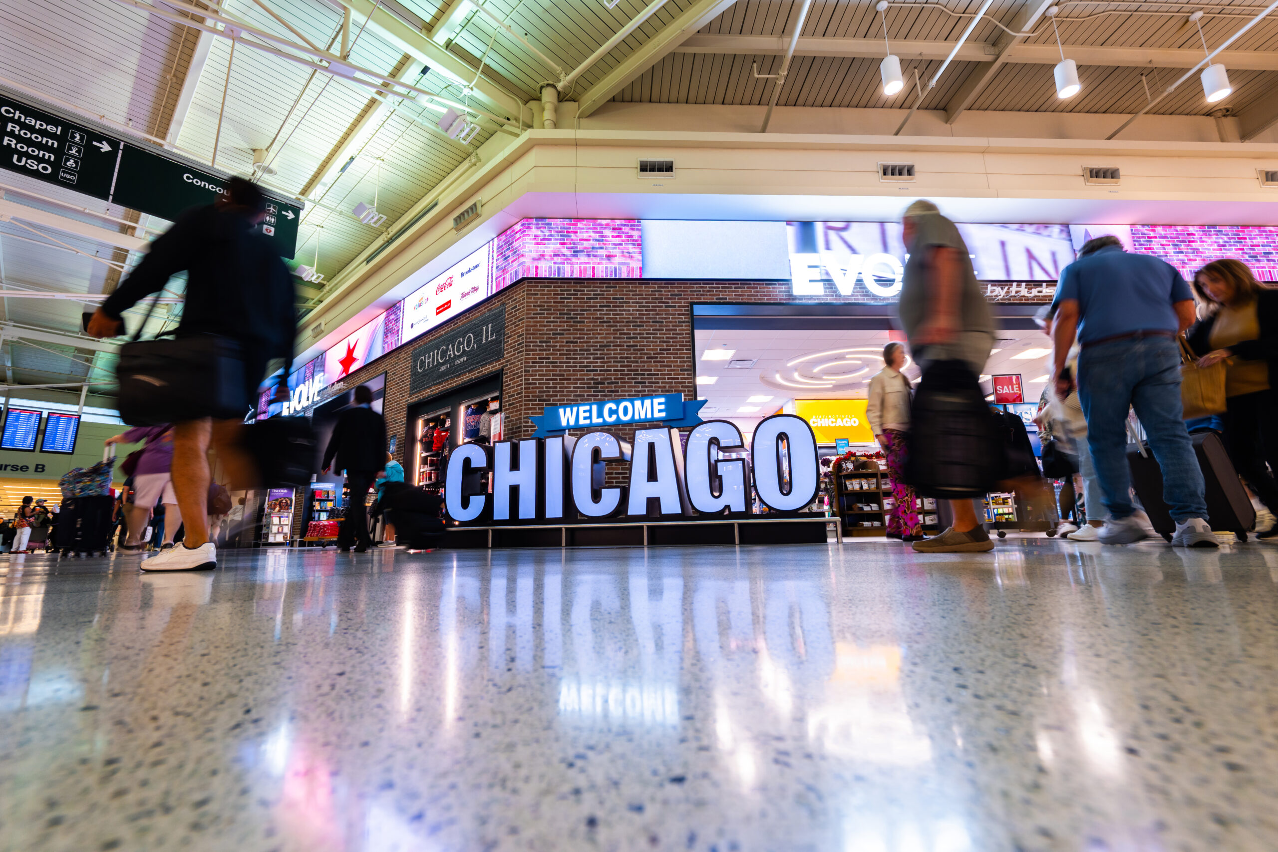 Low-angle view of large illuminated "WELCOME CHICAGO" letters in front of an Evolve by Hudson store with a brick front and a "Chicago, IL" plaque. Colorful digital banners wrap the top of the store. Blurred travelers with bags pass by, and the letters reflect in the polished terrazzo floor.