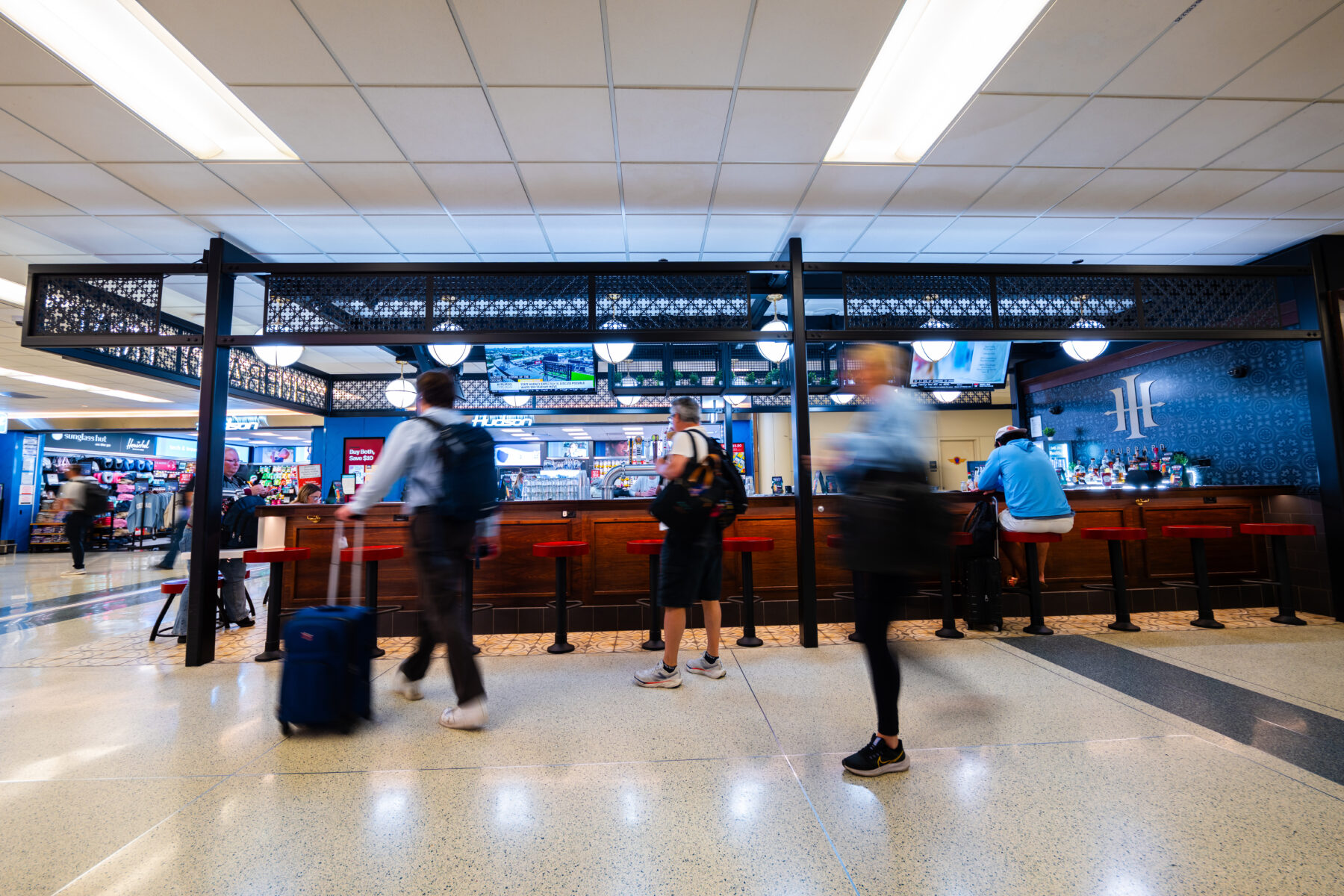 Hubbard Inn bar in an airport concourse, with a wood-paneled counter, red-topped stools and a black metal frame with decorative lattice panels and globe pendant lights. A blue patterned tile wall shows the bar's "HI" logo. Blurred travelers with rolling luggage pass in front, and a Hudson store and Sunglass Hut are visible in the background.