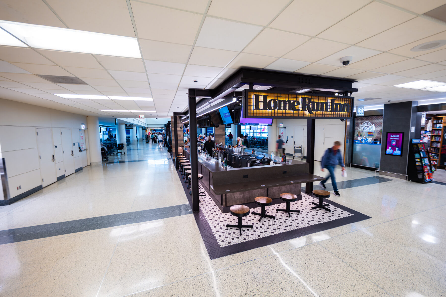 Home Run Inn bar kiosk with an illuminated sign, a black steel frame and wood-topped stools on a black-and-white hexagon tile floor. It sits in a bright terrazzo concourse near gates B16 and B17. A bookstore is on the right, and travelers walk toward the gates in the distance.